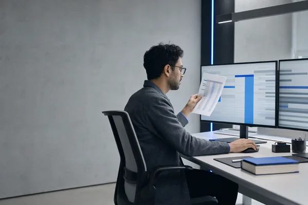 A finance clerk seen from the side at a desk with two monitors, one showing an abstract blue-and-grey ledger, a hand resting on the mouse and a printed document held against the screen for comparison, cool office light