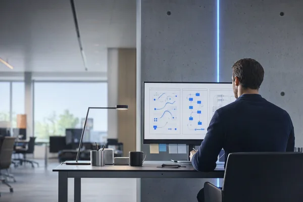 An operations lead seen from behind at a desk with a large monitor showing three abstract blue process flows side by side, sticky notes along the desk edge and a quiet modern office beyond
