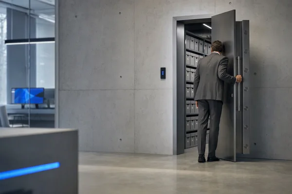 A person in a grey suit seen from behind closing the heavy steel door of a records room lined with labelled grey boxes, a small blue-lit access panel on the wall, cool even light