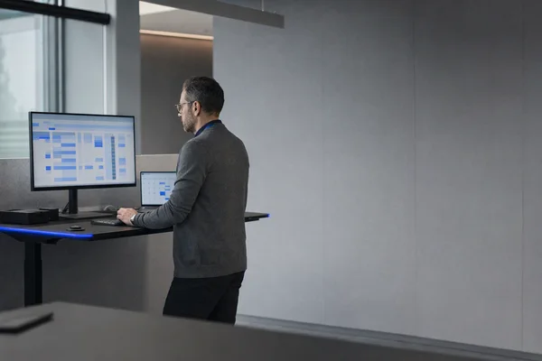 A finance controller seen from the side at a standing desk in an open office, a wide monitor showing an abstract blue-and-grey ledger with a column of version numbers, a second screen with a calendar, grey walls and a thin blue LED strip under the desk
