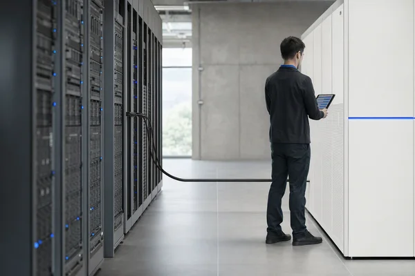 A server room corridor seen along its length, two racks of older grey equipment on the left and one new white rack on the right joined by a single bundled cable, a technician from behind checking a handheld screen with abstract blue-and-grey rows, thin blue status lights