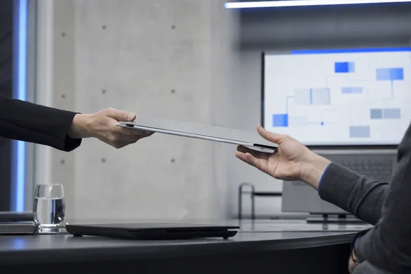 Close view of one colleague handing a document folder to another across a desk, a laptop with an abstract blue process diagram softly out of focus behind, clean office light