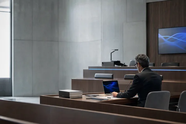 An empty modern hearing room with wooden benches, a long table, a laptop and stacked documents