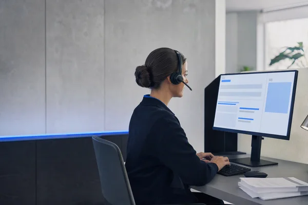 A customer service employee with a headset seen from the side at a tidy desk, reading a drafted reply on a monitor of abstract blue text blocks, a short stack of paper invoices beside the keyboard, soft daylight
