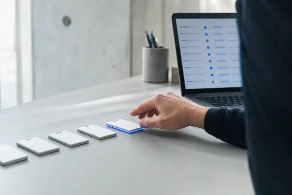 A row of identical white access badges on a desk in a clean office, one of them lit by a thin blue line along its edge, an administrator's hand reaching for it, a laptop in the background showing an abstract blue-and-grey list, shallow depth of field