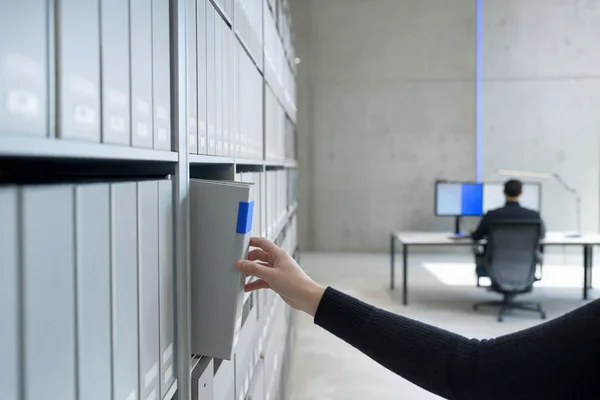 Tidy grey archive shelves of labelled binders in a modern records room, one binder pulled halfway out with a blue tab and a hand reaching for it, shallow depth of field