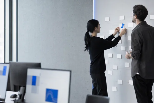 Two people seen from the side rearranging white cards on a large wall board of connected boxes in a bright workspace, one new card marked with a strip of blue tape