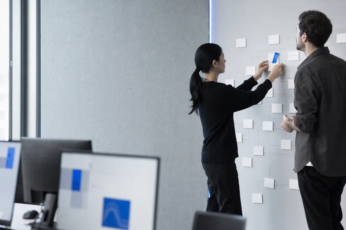 Two people seen from the side rearranging white cards on a large wall board of connected boxes in a bright workspace, one new card marked with a strip of blue tape