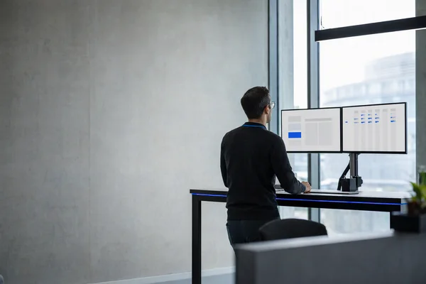 A logistics office seen from behind an operator at a standing desk: on the left monitor a single abstract blue-and-grey form, on the right a wide abstract table of thin columns, a thin blue LED strip along the desk edge, morning light from a tall window