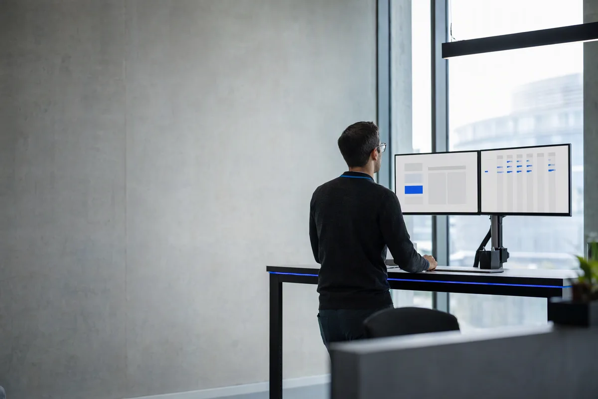 A logistics office seen from behind an operator at a standing desk: on the left monitor a single abstract blue-and-grey form, on the right a wide abstract table of thin columns, a thin blue LED strip along the desk edge, morning light from a tall window