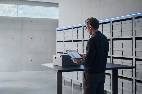 A quiet archive room seen from the side: an archivist at a standing desk with a single tablet showing an abstract blue-and-grey ledger, behind him long rows of identical grey storage boxes under a thin blue LED line, morning light from a high window