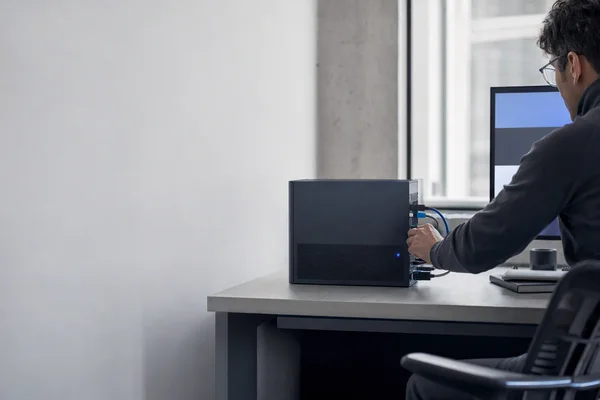 A compact graphite server on a desk in the corner of a small, clean office, a single blue status light glowing, an engineer's hand adjusting a cable, soft daylight from a window behind