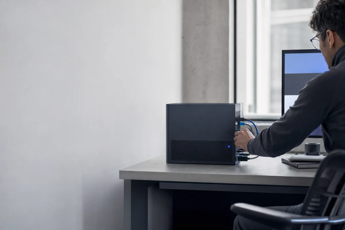 A compact graphite server on a desk in the corner of a small, clean office, a single blue status light glowing, an engineer's hand adjusting a cable, soft daylight from a window behind