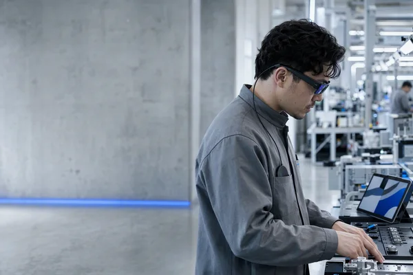 A frontline worker in a bright assembly hall seen from the side, wearing slim unbranded display glasses connected by a thin cable to a small compute unit in the chest pocket of a grey work jacket, looking down at a workbench with parts and a tablet, a faint blue reflection on the lenses