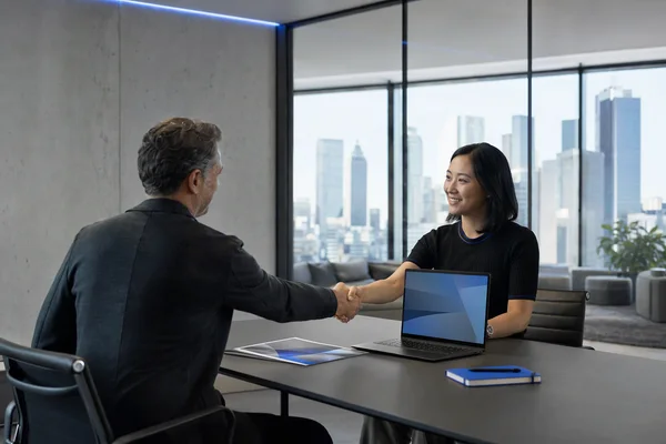 Two people shaking hands across a meeting table with a laptop and a printed proposal