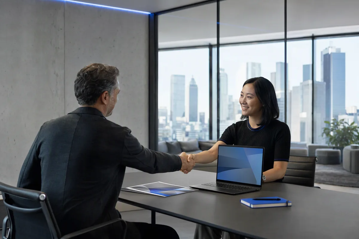 Two people shaking hands across a meeting table with a laptop and a printed proposal