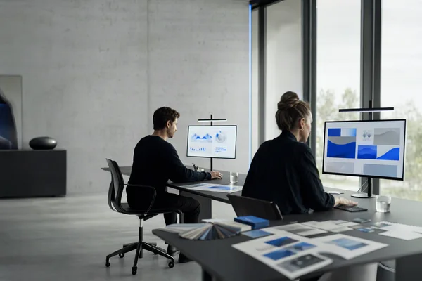 Two people at a long table with large monitors, printed proofs and colour swatches in a bright studio