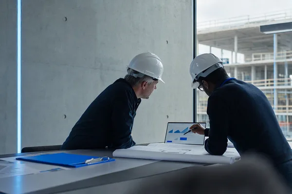 Engineers in hard hats reviewing a printed plan and a tablet in a construction site office