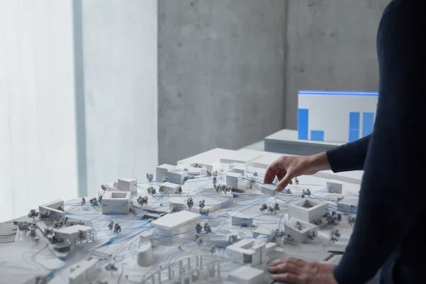 A hand placing a block on a white architectural model of a campus, with threads linking the buildings